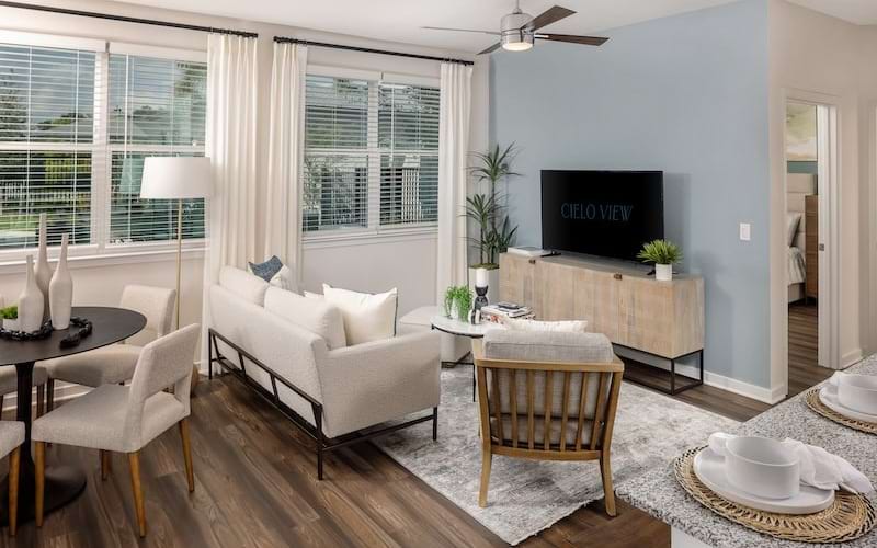 Kissimmee, FL, Luxury Apartments with In-Unit Washer and Dryer - Cielo View - Bright Living Room with Sofa, Dining Table, Large Windows with Curtains, Wall-Mounted Television, and Media Console.